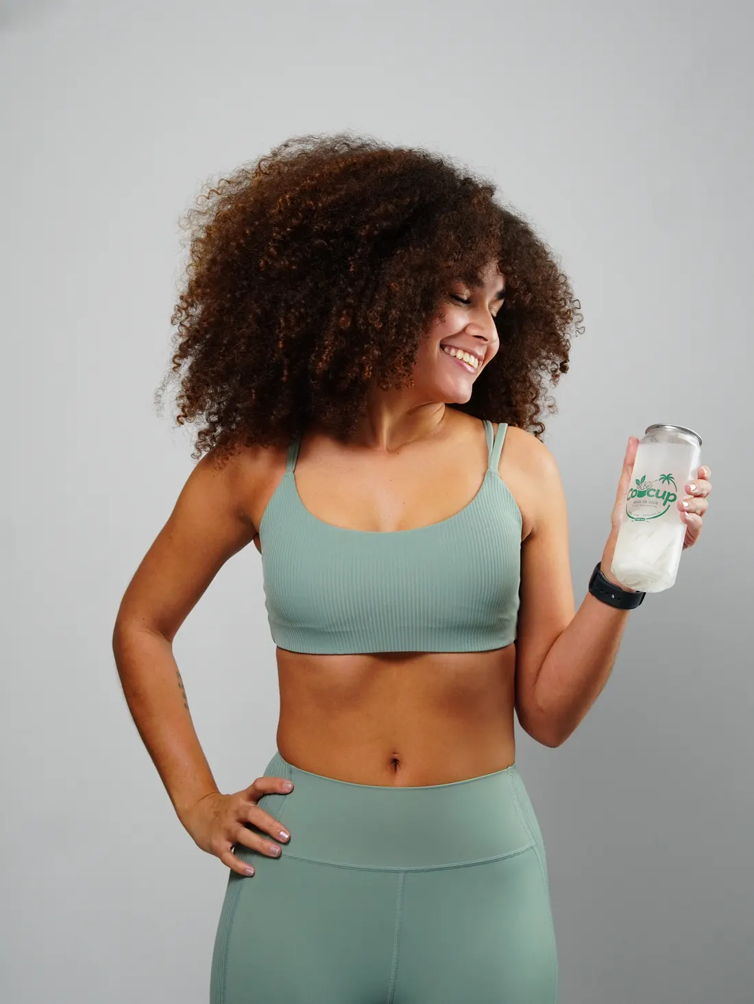 Mulher rindo, de top e legging verde-sálvia, com um copo de Cocup na mão
